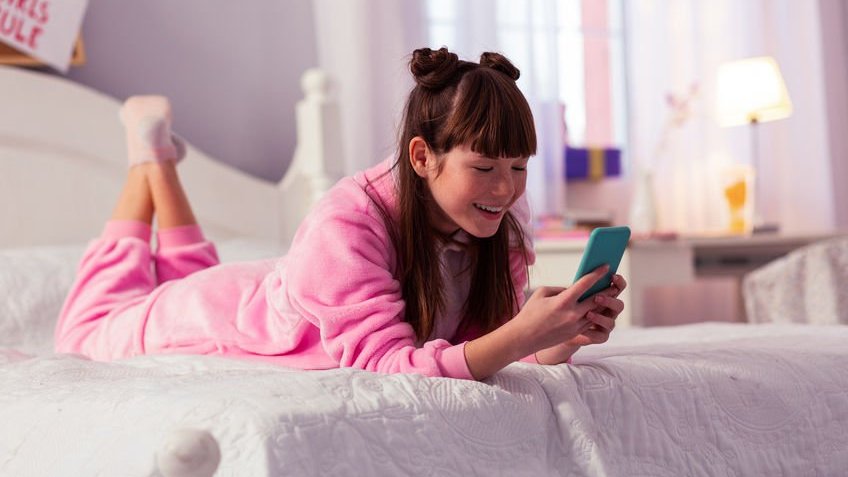 Garota deitada na cama de barriga para baixo usando pijama e mexendo no celular