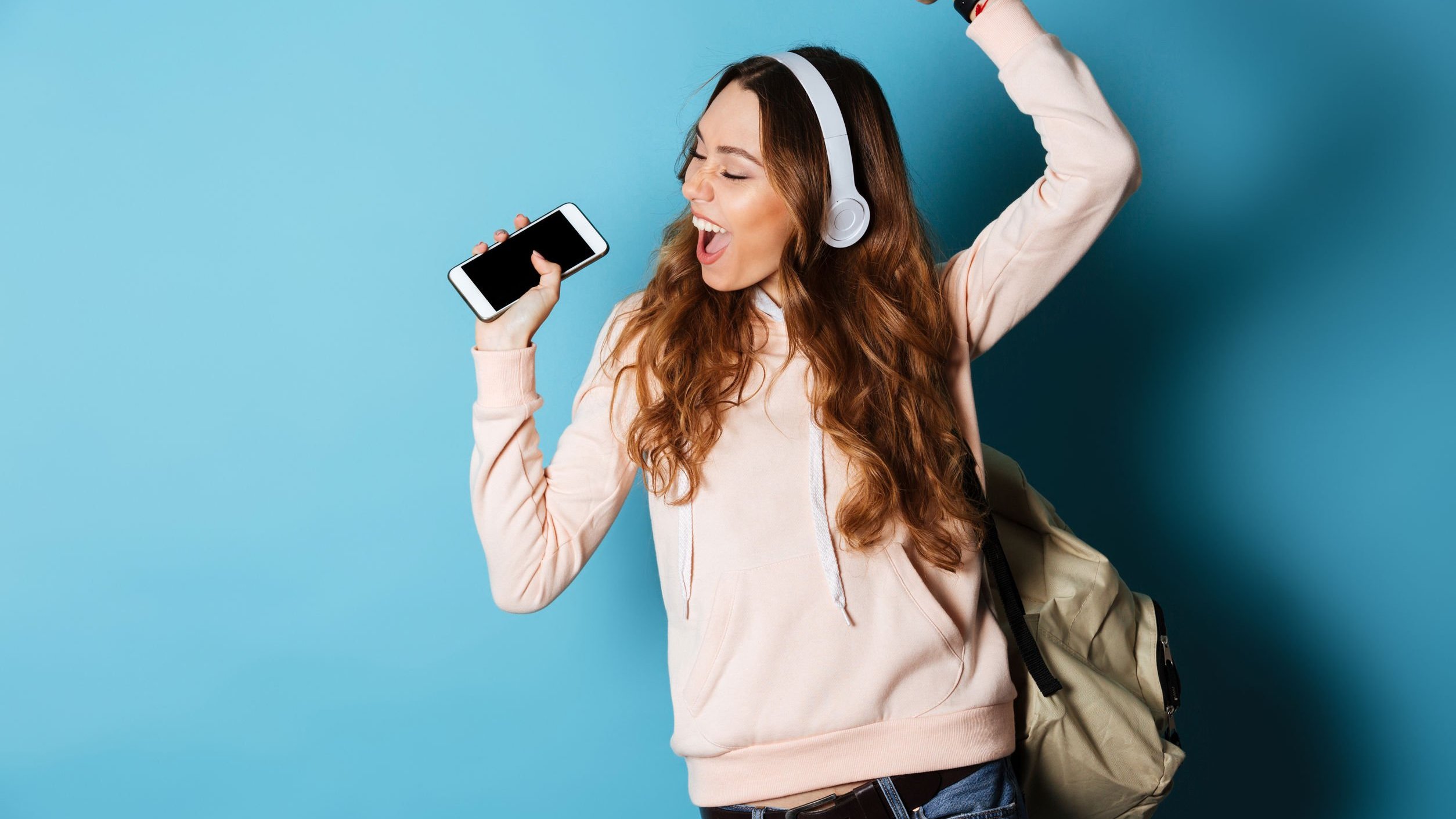 Mulher usando fones, ouvindo música, segurando celular e cantando com a mão pra cima.