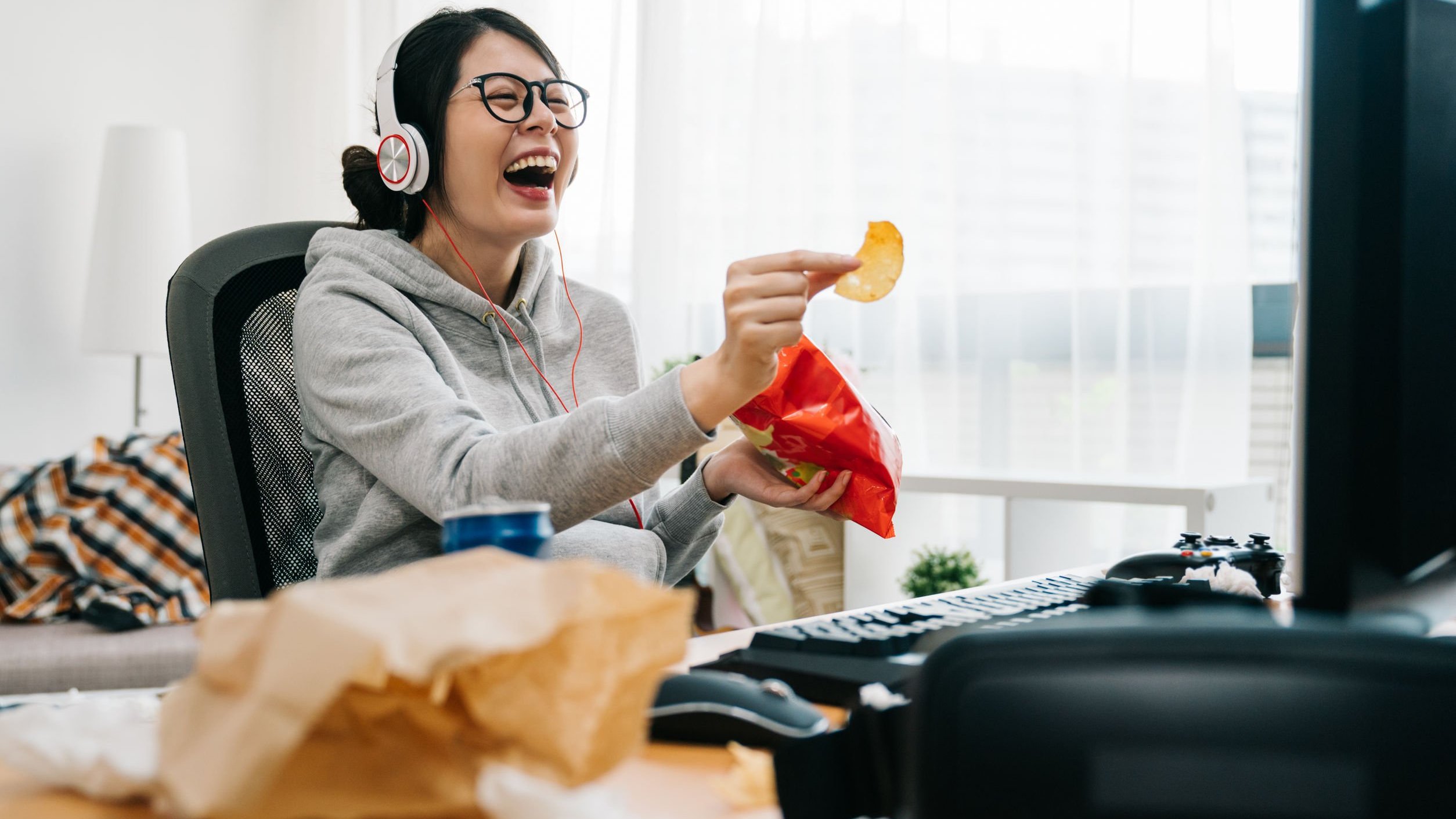 Mulher dando risada enquanto come salgadinho em frente de computador