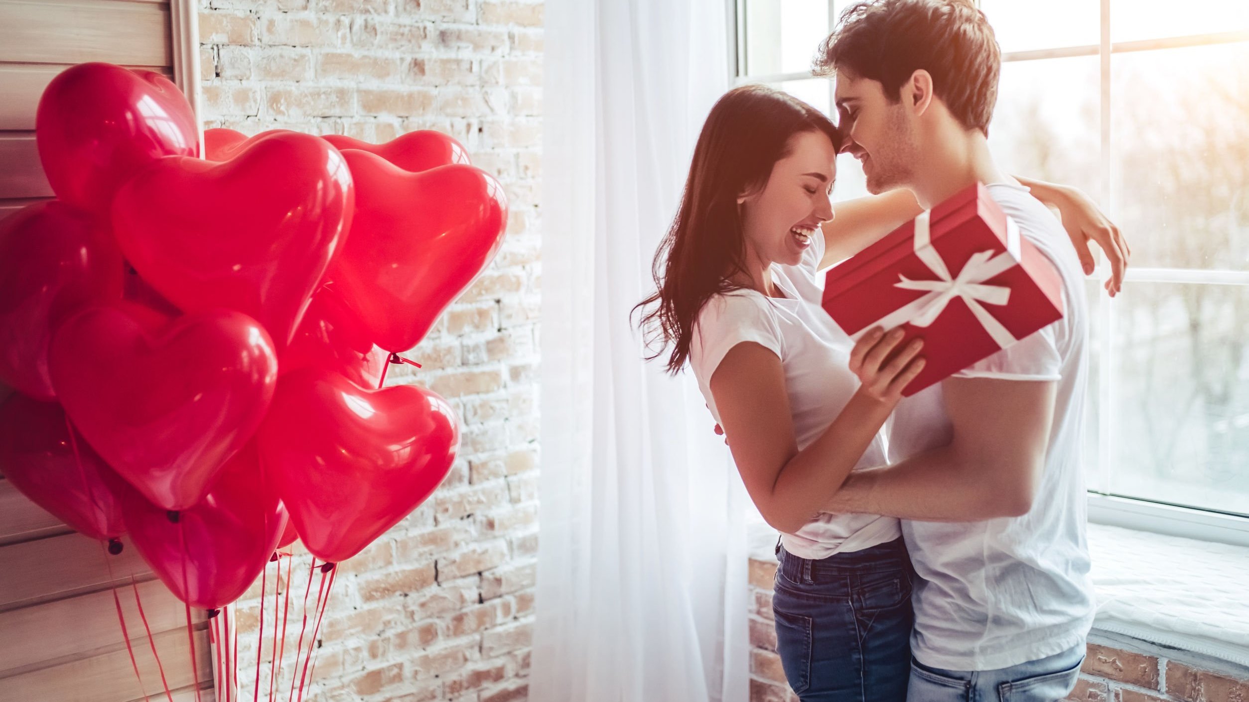 Homem e mulher abraçados e segurando uma caixa de presente, ao lado de balões em forma de coração.