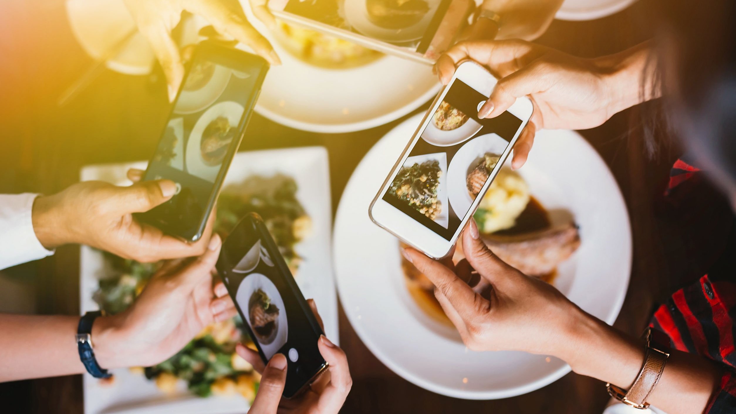 Mãos segurando celulares com câmeras ligadas apontando para a comida na mesa.