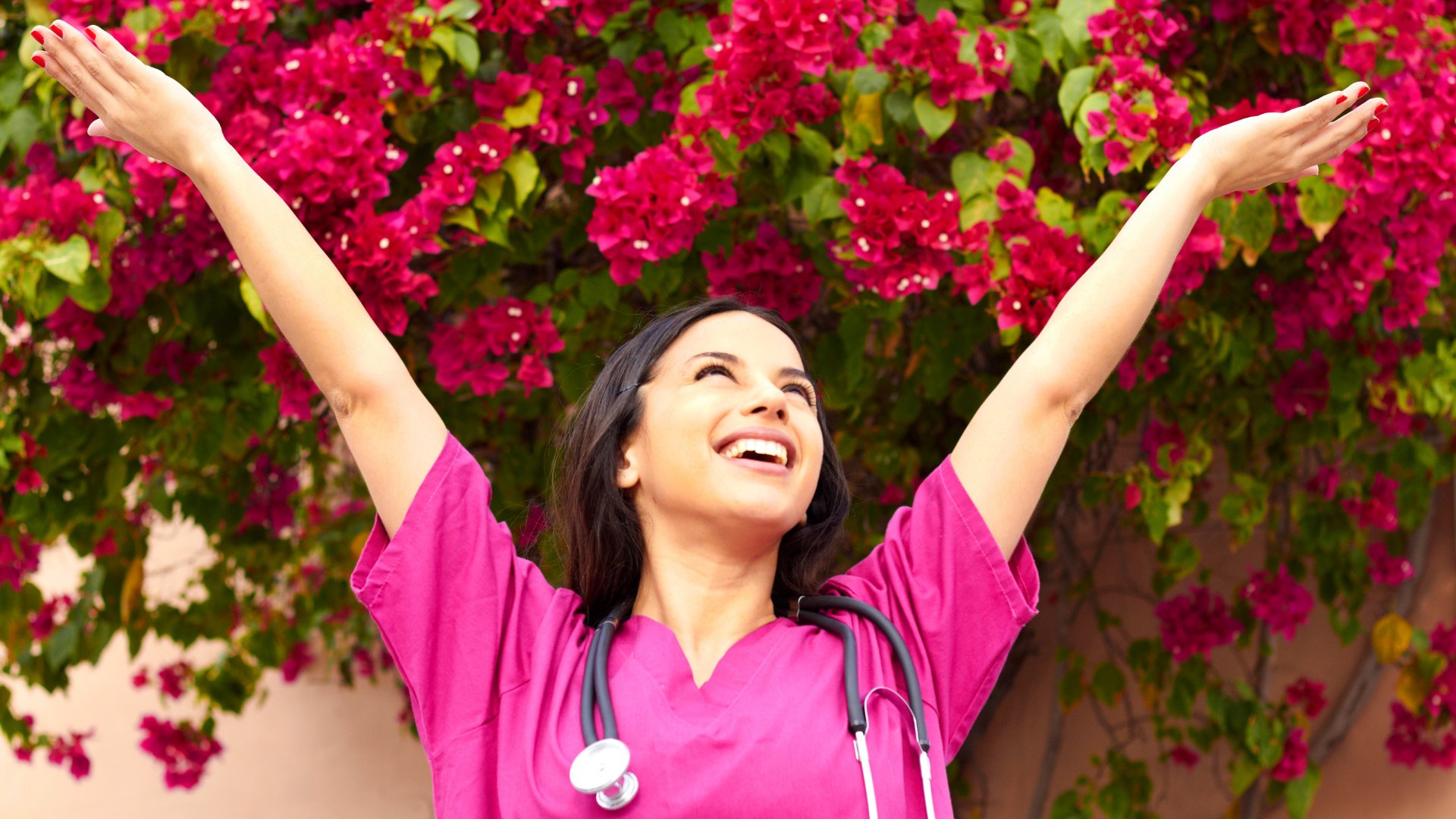 Enfermeira sorrindo de braços erguidos com flores rosas ao fundo