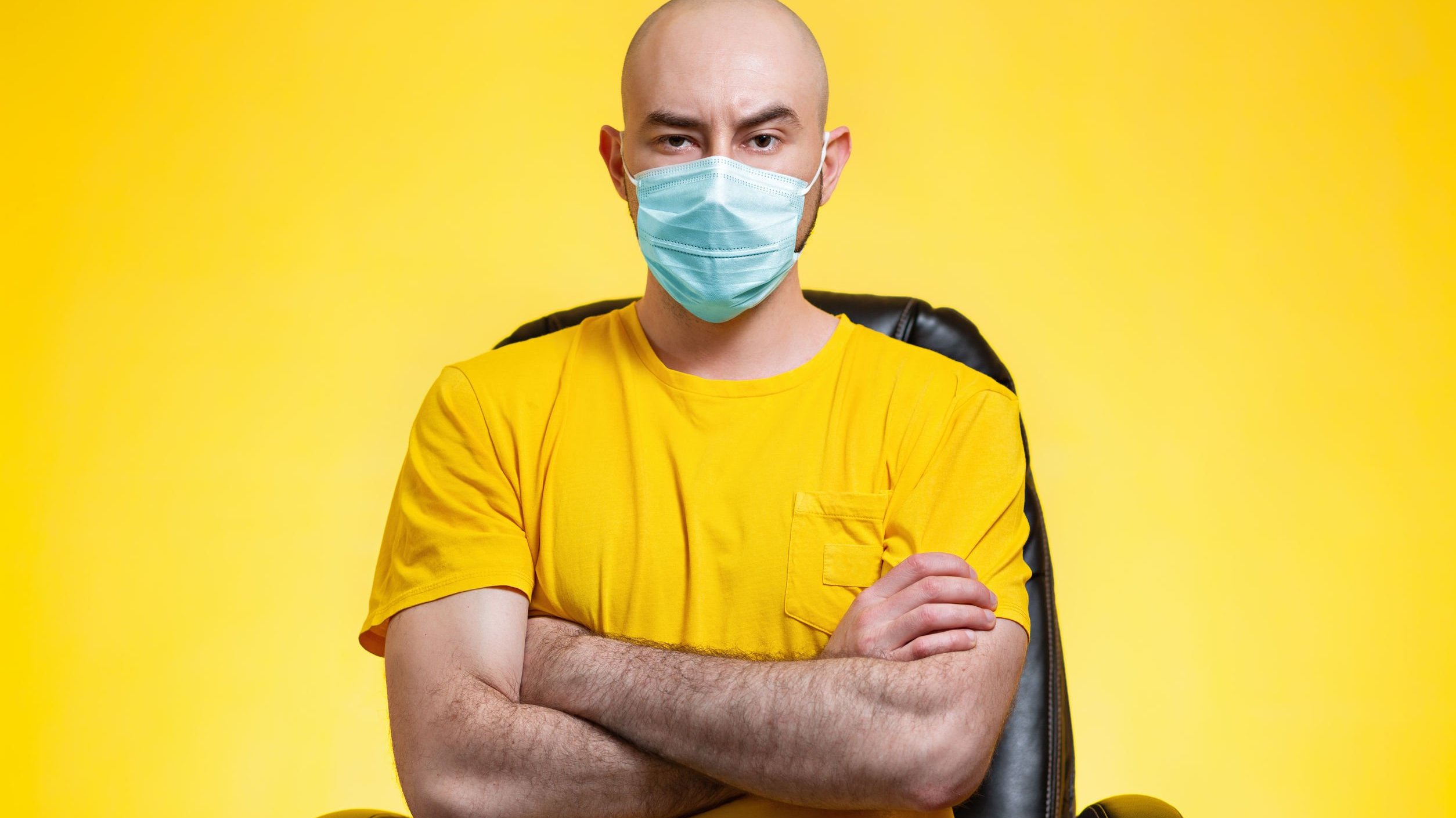 Homem sentado de braços cruzados e uma sobrancelha levantada enquanto usa máscara de proteção
