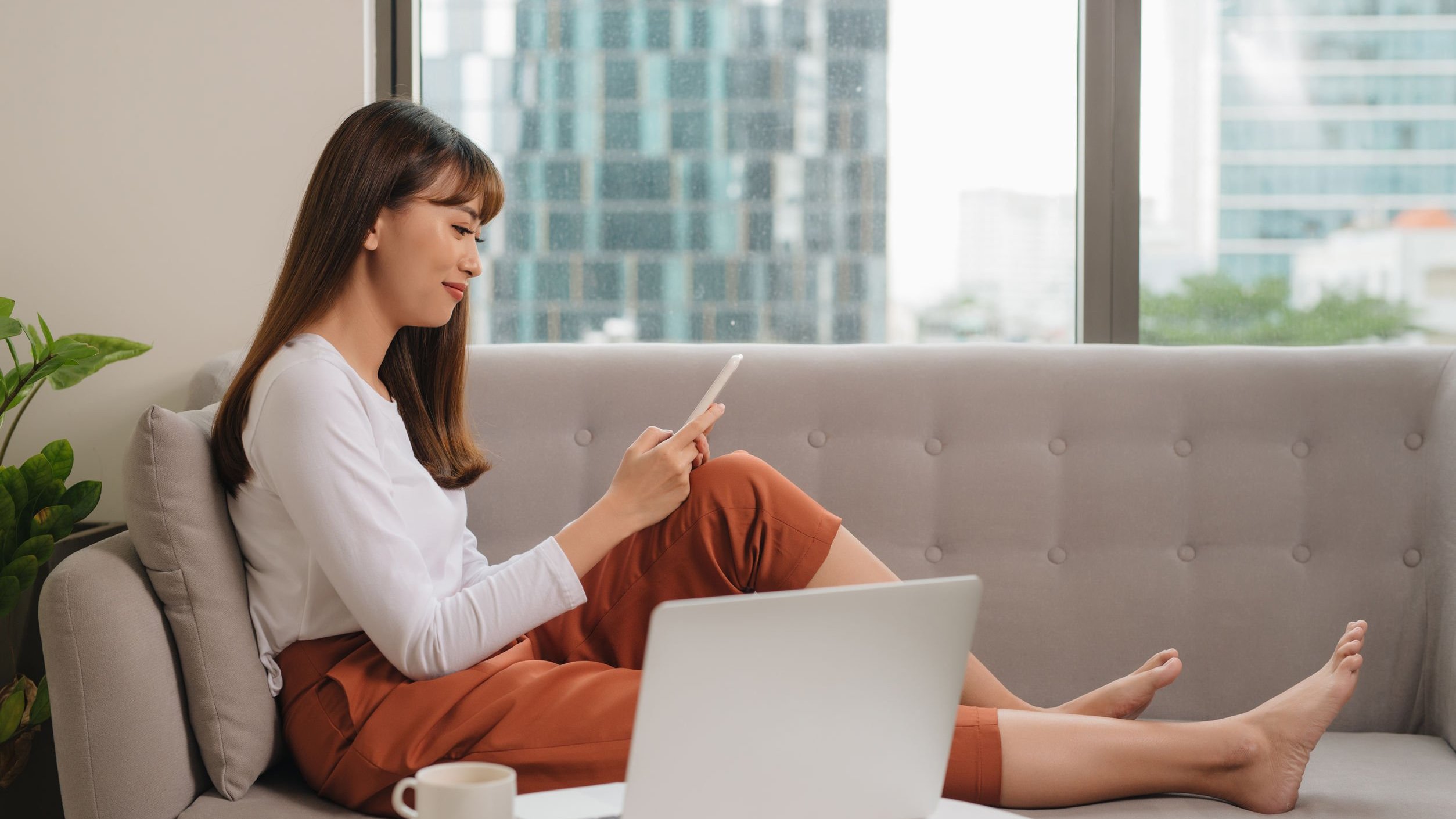 Mulher sentada no sofá ao lado de um notebook e uma caneca, usando seu telefone celular.