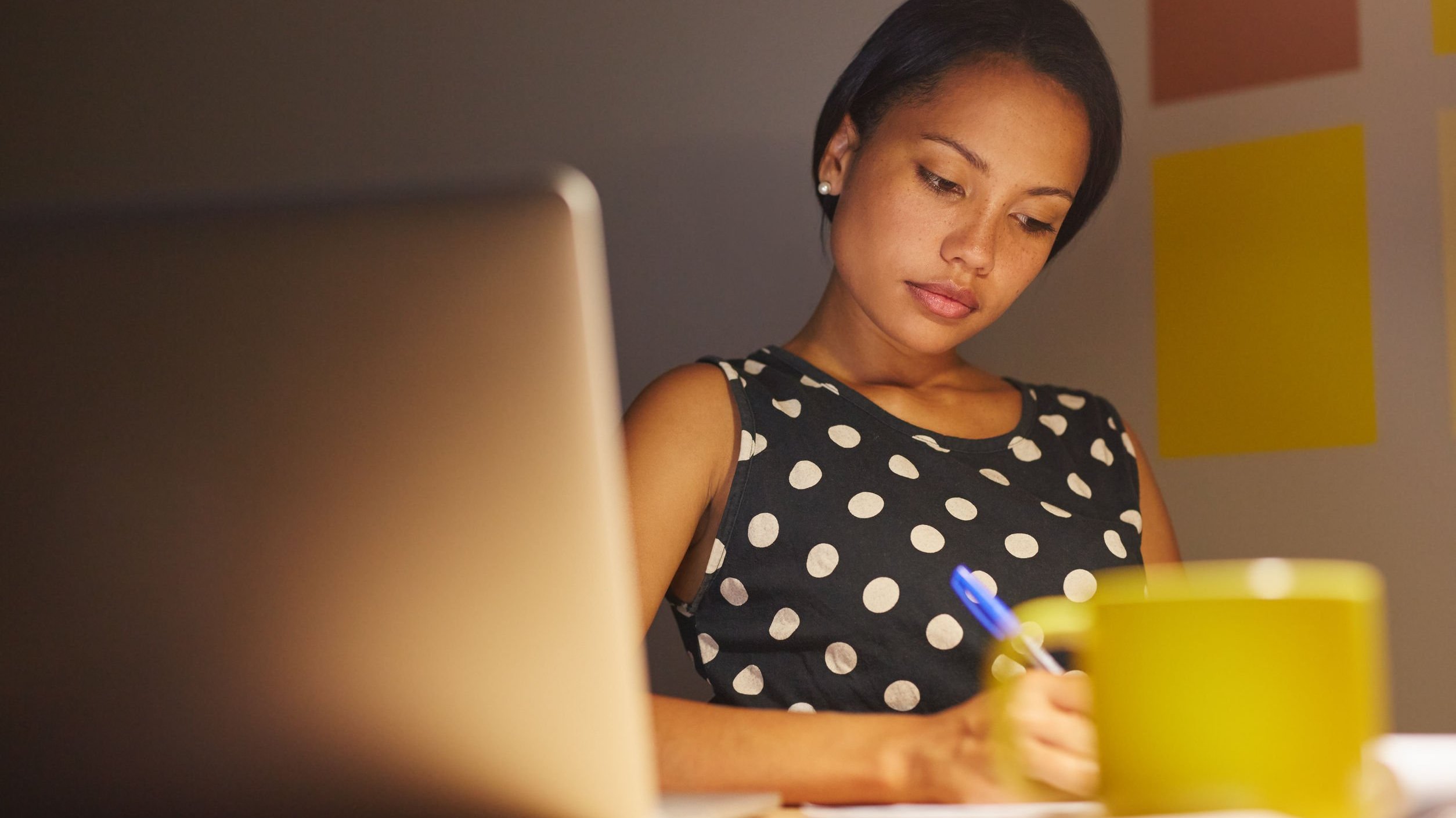 Mulher em seu escritório fazendo anotações em uma folha com seu notebook aberto ao lado