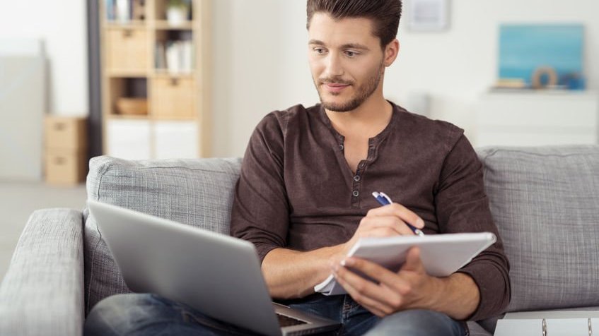 Garoto estudando no sofá de casa com caderno e notebook