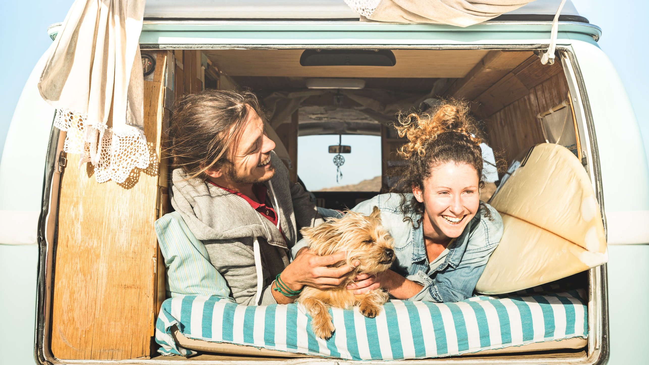 Casal branco deitado dentro de uma Kombi com um cachorro, expressões sorridentes