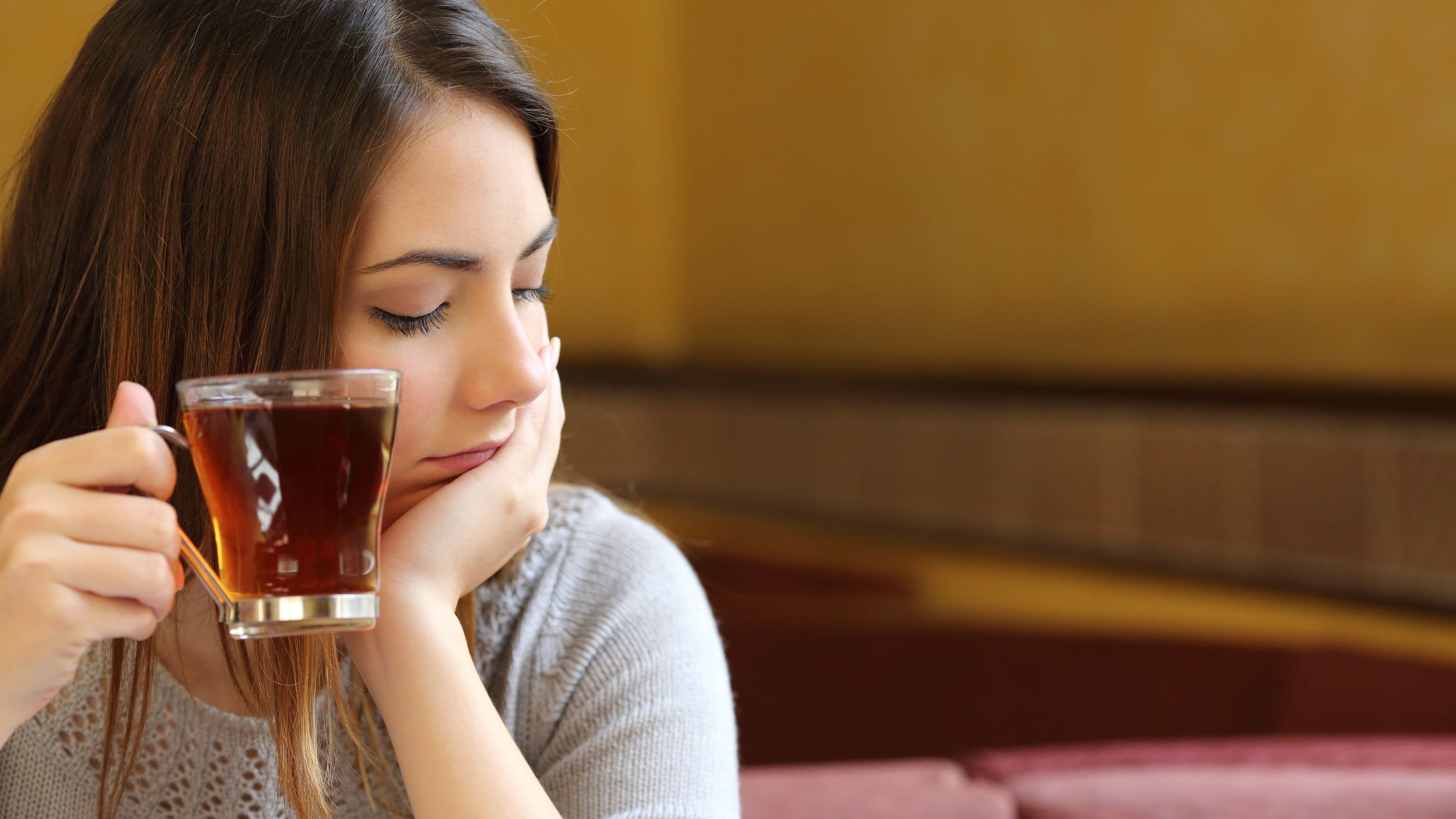 Mulher branca de cabelos castanhos e lisos, com expressão chateada, segurando xícara de café com uma mão e a outra apoiada no queixo