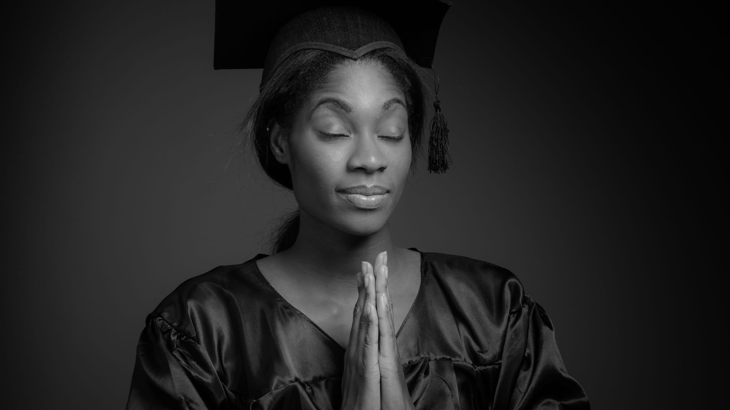 Foto preta e branca de mulher negra com roupas de formatura, orando de olhos fechados