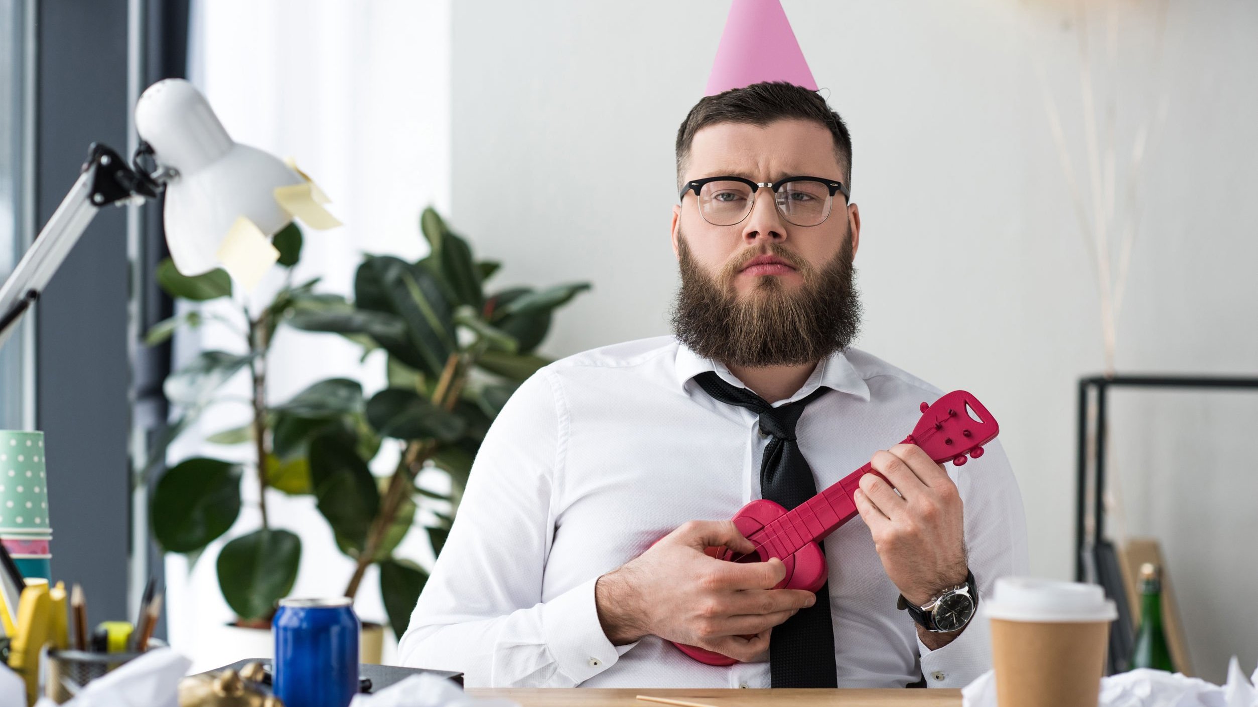 Homem usando chapeuzinho de aniversário e segurando cavaquinho, sentado em escritório.