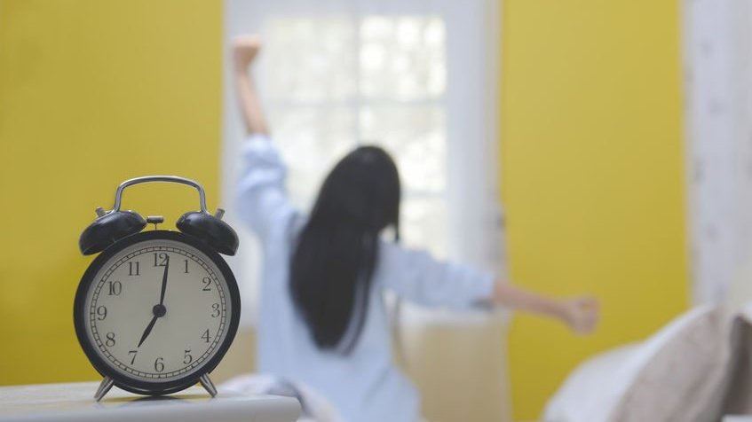 Mulher sentada na cama se espreguiçando ao lado de um relógio, que marca as 7 horas