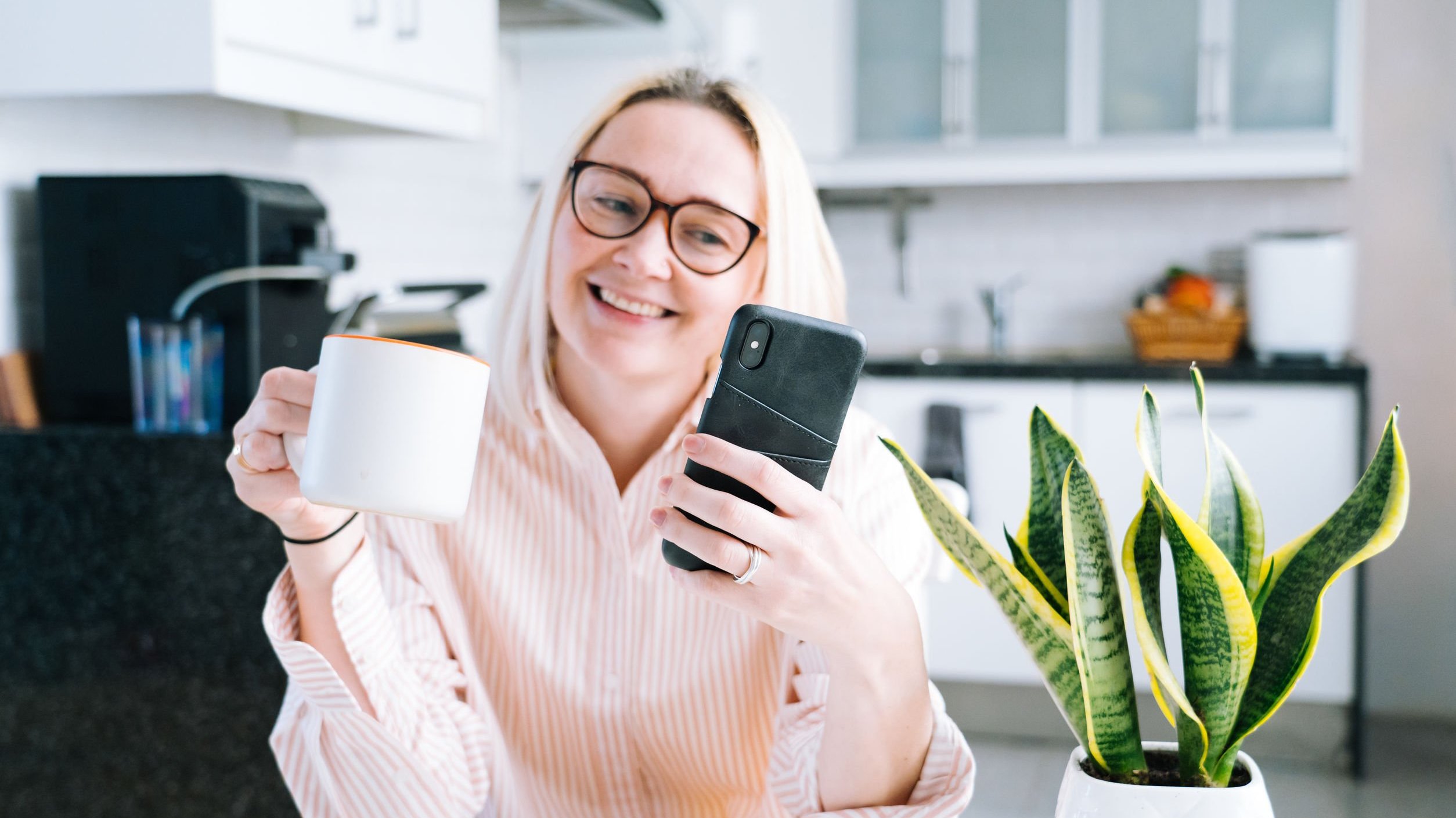 Mulher branca e loira de óculos preto, segurando uma caneca e um celular, com expressão sorridente