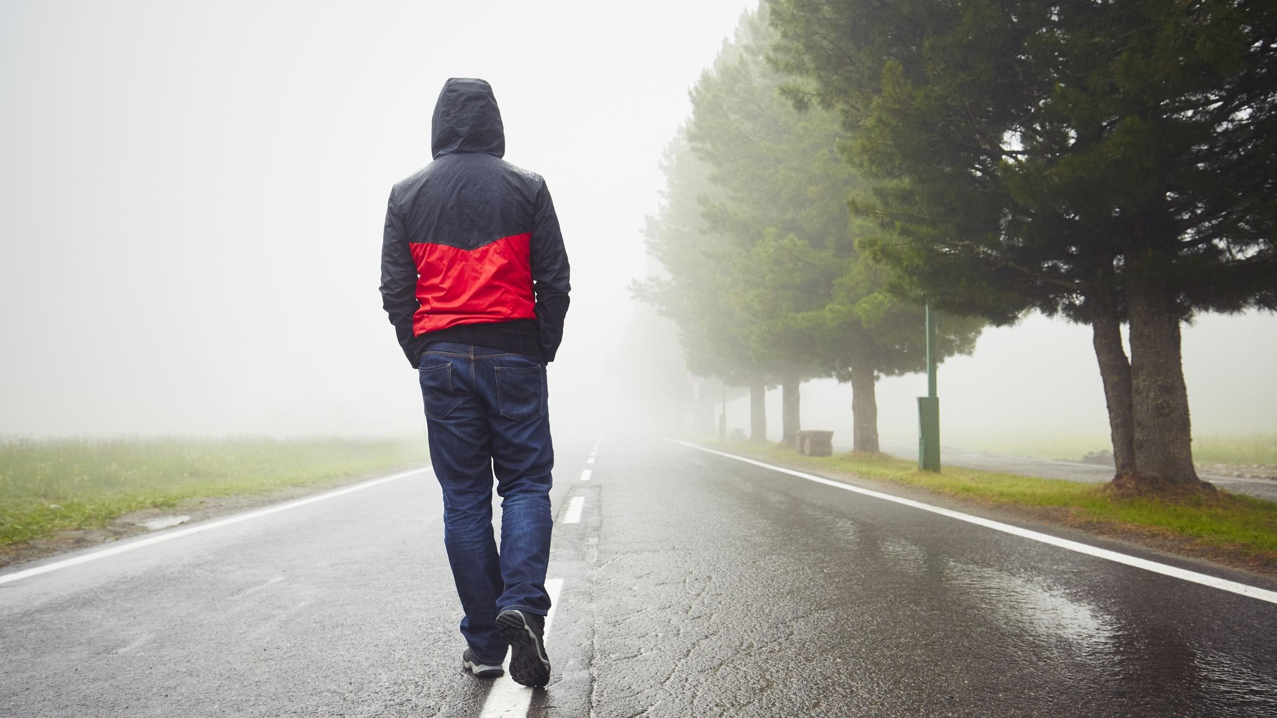 Homem de costas andando numa rua meio à neblina e chuva, com capuz na cabeça e mãos no bolso.
