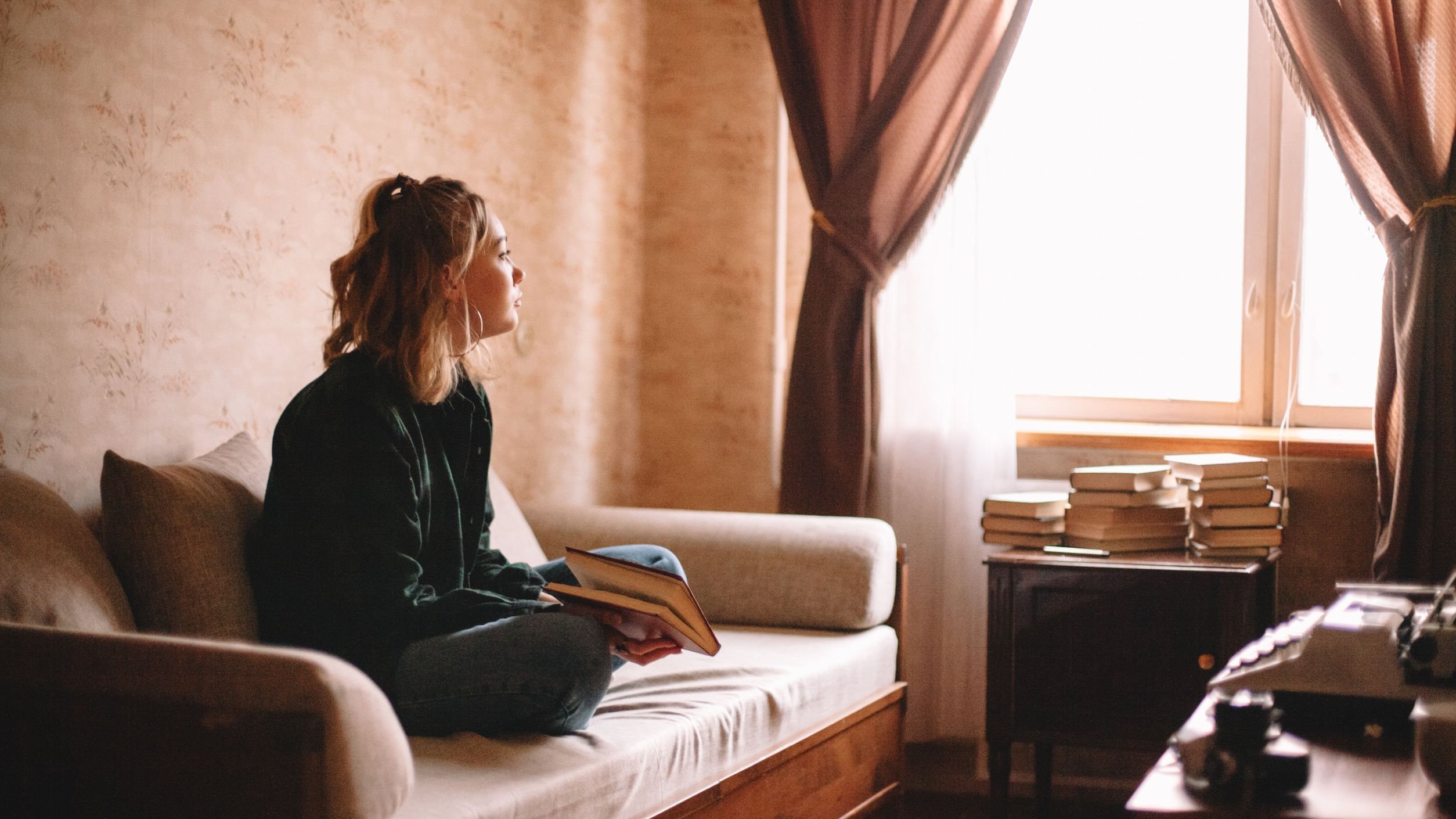 Menina adolescente sentada no sofá de sua casa, com um livro em mãos, olhando pela janela