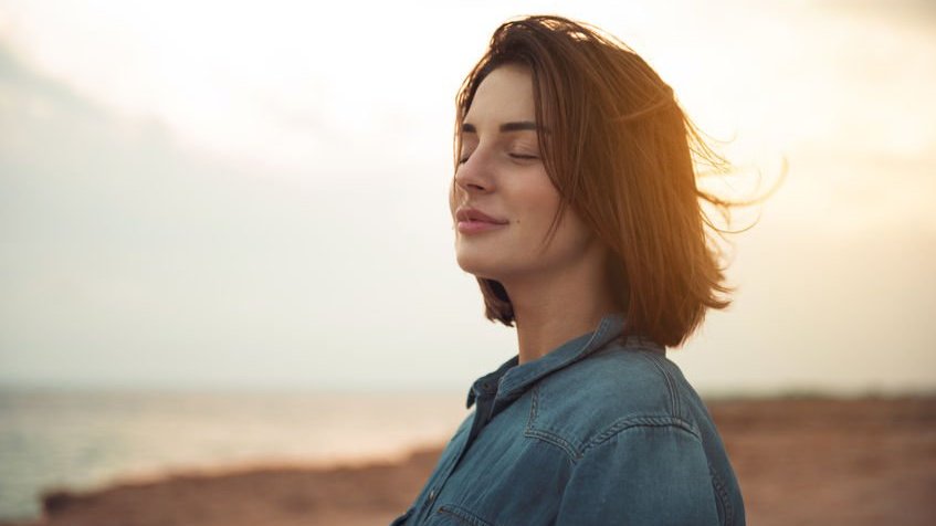 Mulher de olhos fechados e vento em seus cabelos com sol refletindo ao fundo