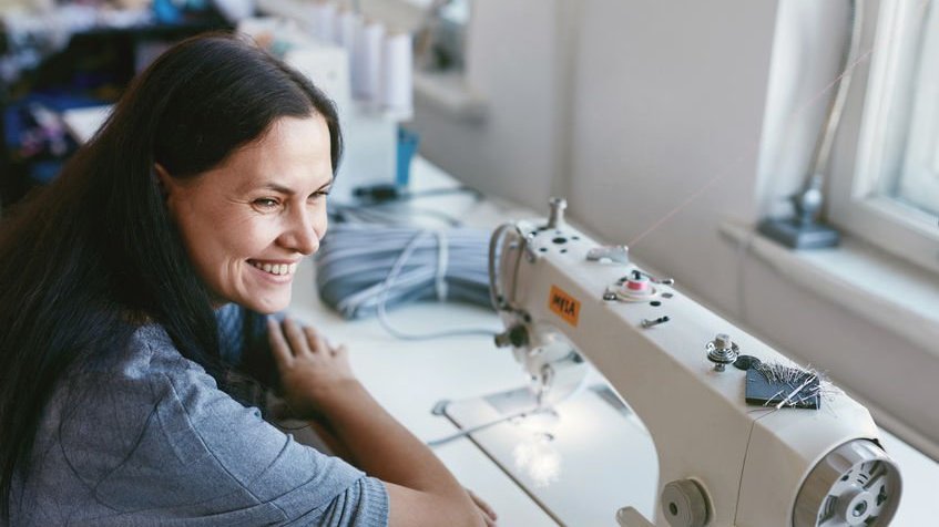 Mulher sorrindo sentada em mesa com máquina de costura em frente