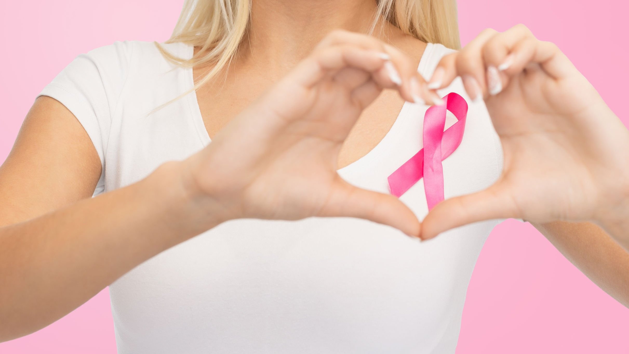 jovem com uma camiseta branca, lisa, e uma fita de conscientização do câncer de mama, mostrando o formato de um coração sobre um fundo rosa