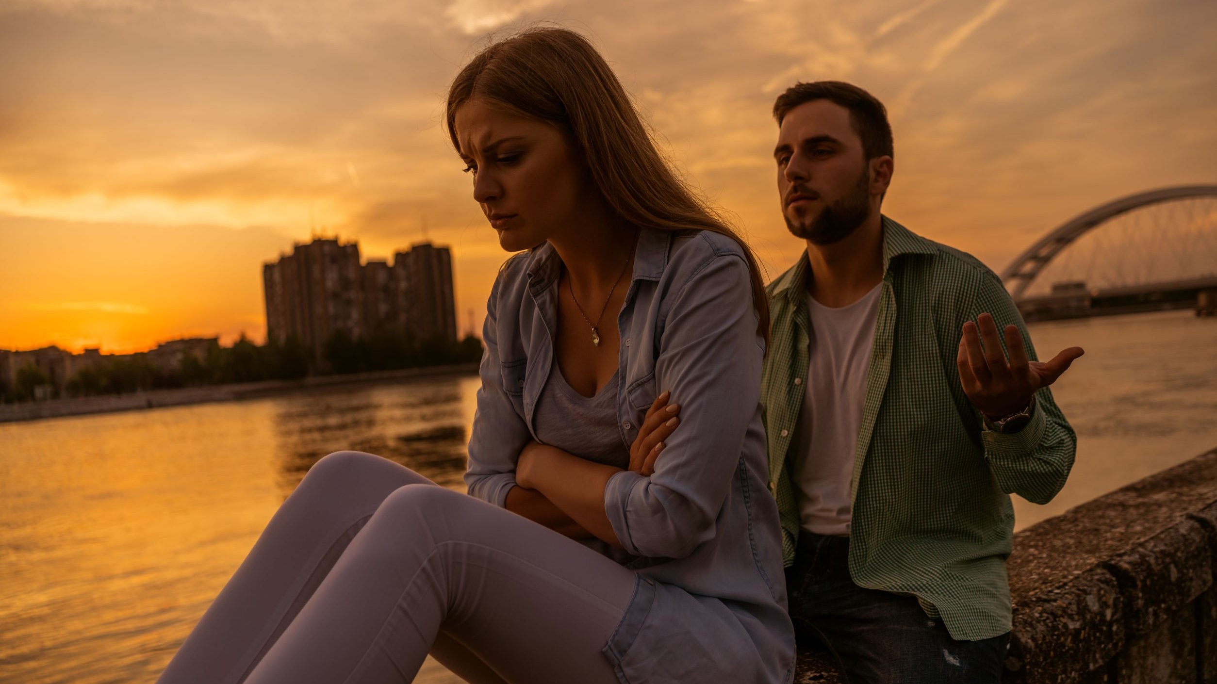 Homem e mulher brancos sentados numa mureta ao lado de um rio. Mulher com braços cruzados e expressão triste, homem com braços abertos e expressão de súplica.