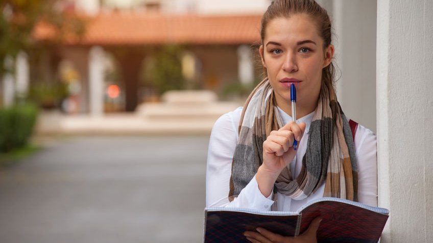 Mulher com caneta posicionada nos lábios e segurando caderno