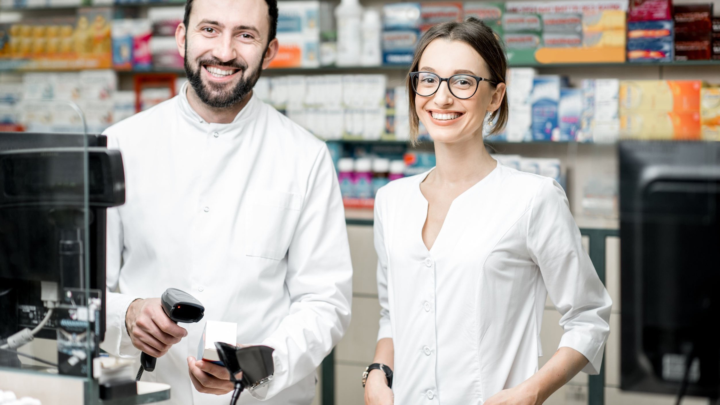 Homem e mulher, ambos farmacêuticos, sorrindo atrás de balcão de farmácia.