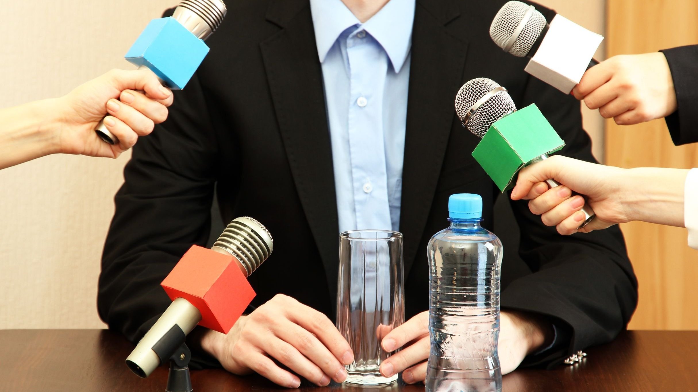 Homem dando entrevista, cercado de mãos segurando microfones.