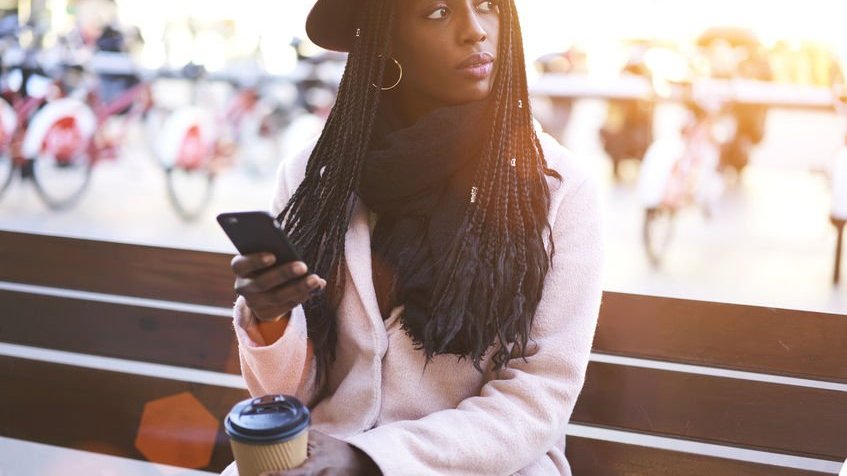 Mulher sentada em banco mexendo no celular com copo de café usando chapéu