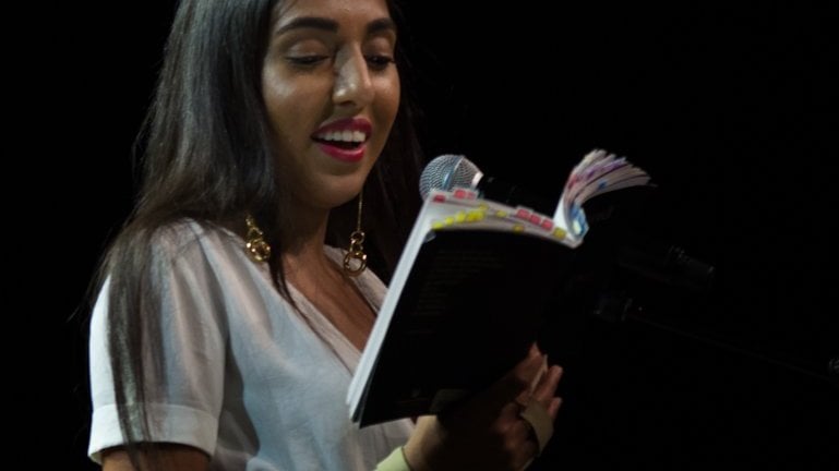 Rupi Kaur lendo livro e segurando microfone