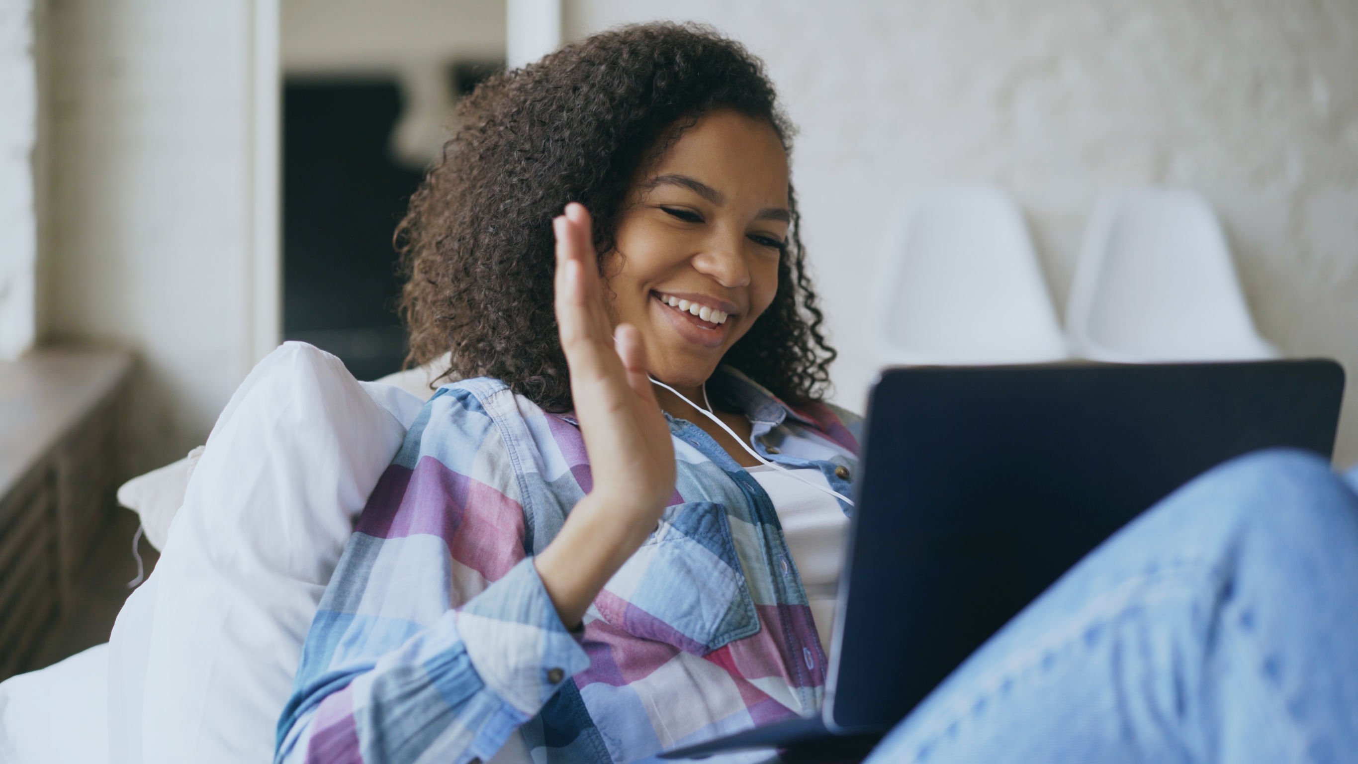 Garota deitada em uma cama, sorrindo e acenando enquanto conversa via videoconferência em um laptop com os amigos.