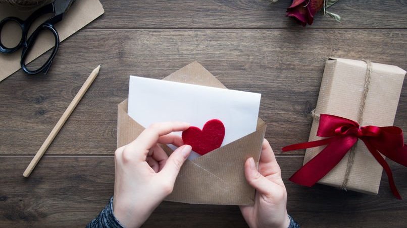 Par de mãos guardando uma carta e um pequeno coração vermelho dentro de um envelope marrom