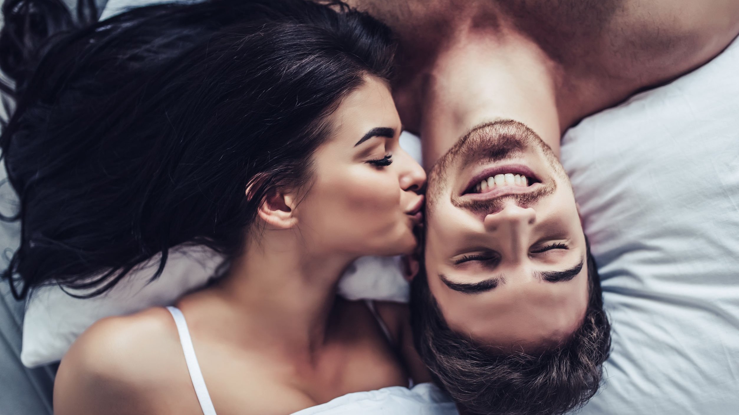 Mulher beija o rosto de um homem, os dois estão deitados em uma cama.