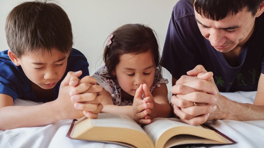 Duas crianças e um adulto ajoelhados rezando debruçados na bíblia