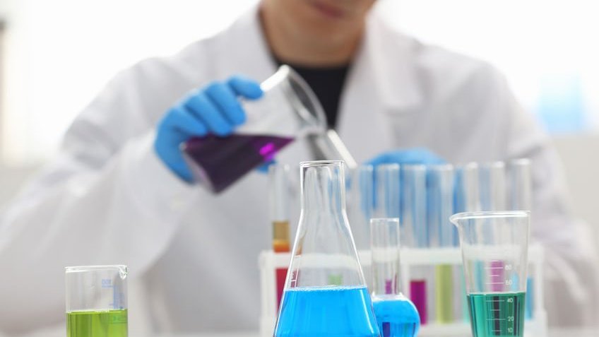 Um químico do sexo masculino segura um tubo de ensaio de vidro em sua mão ao fundo, e na mesa possui vários tubos de ensaio