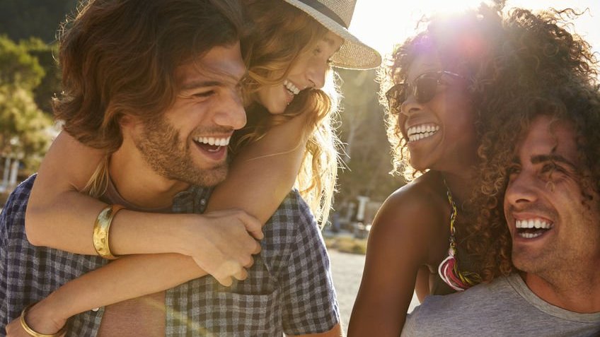 Uma mulher branca está sorrindo para uma mulher negra, enquanto está nas costas de um homem branco alegre. Já a mulher negra está nas costas de um homem negro sorridente , enquanto sorri de volta para a mulher branca