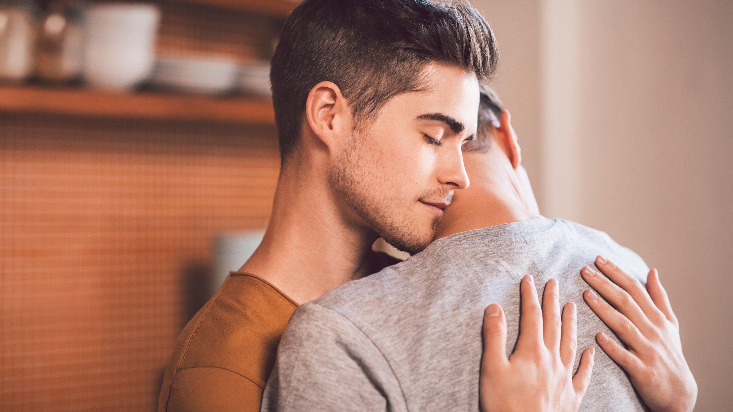 Homem de olhos fechados abraça outro homem, com as mãos posicionadas em suas costas.