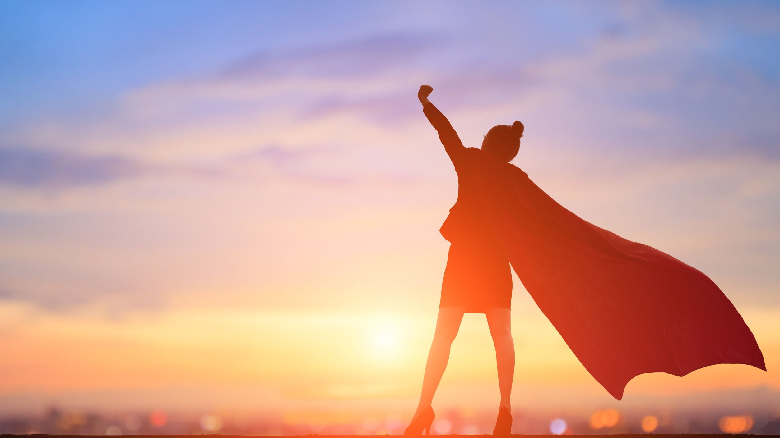 Mulher vestida com uma capa de heroína enquanto ergue um braço direcionado ao céu.