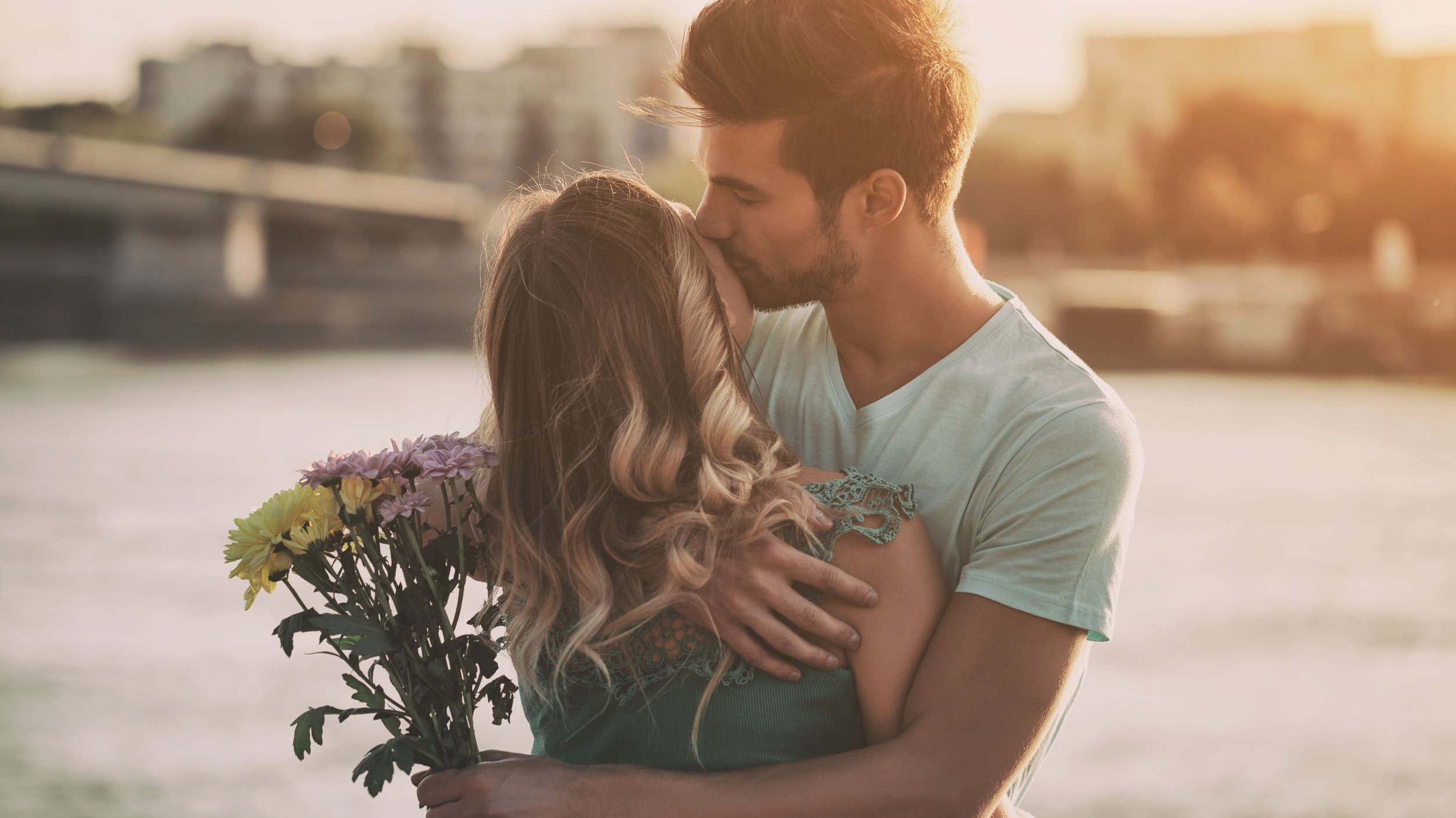 Homem com buquê de flores em uma das mãos abraça mulher enquanto beija o rosto dela.