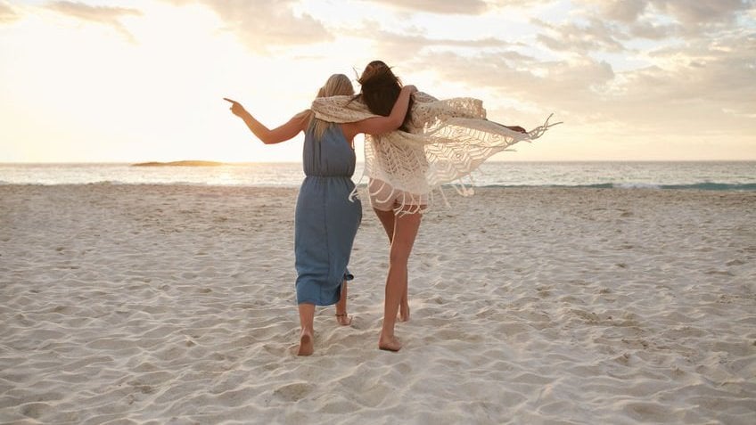 Duas amigas abraçadas andando na areia e indo de encontro ao mar