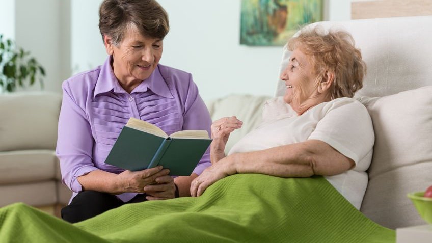 Mulher idosa deitada na cama enquanto outra mulher lê um livro