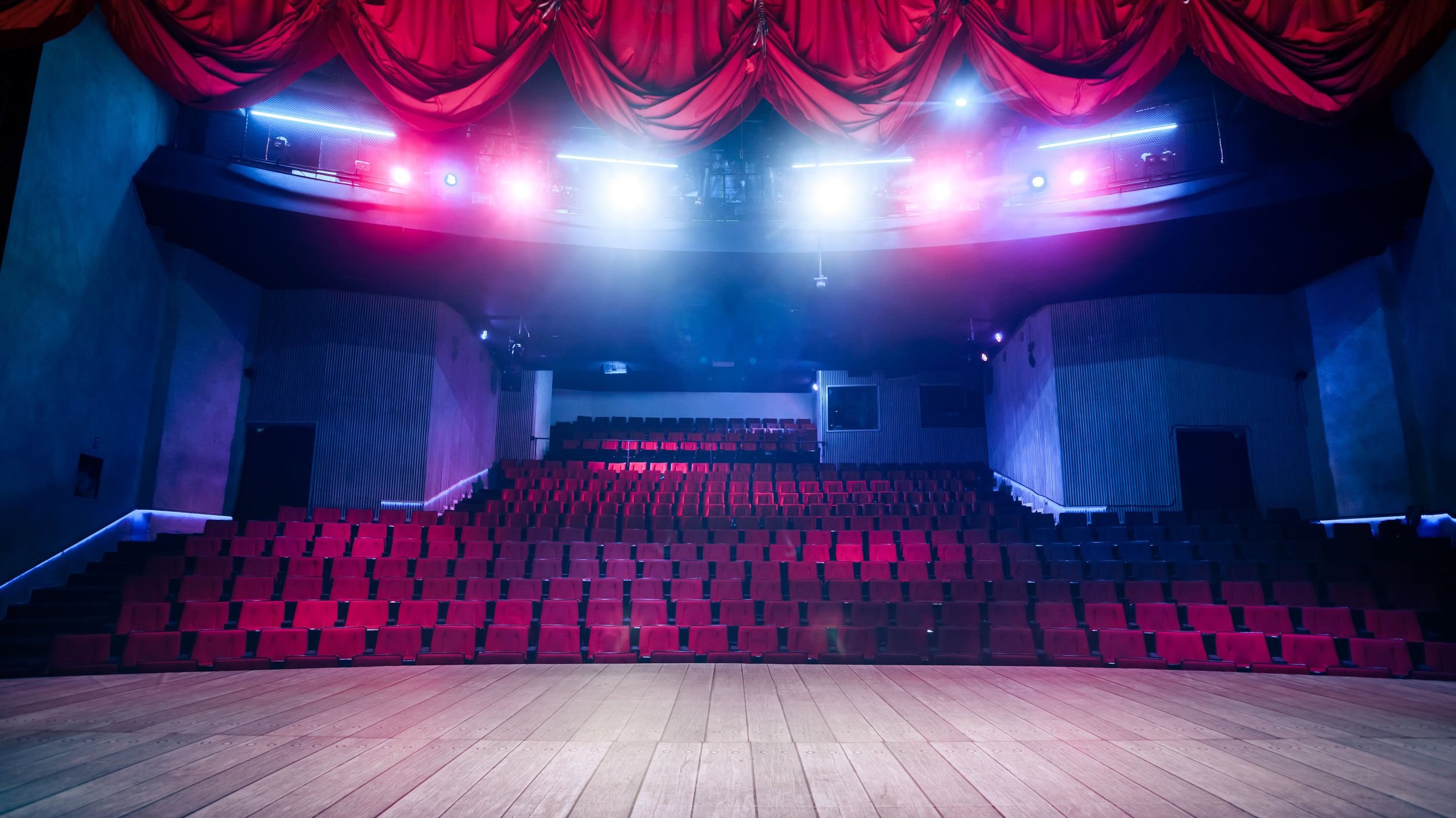 Cortina vermelha e palco de teatro. Ao fundo, assentos iluminados por luzes.