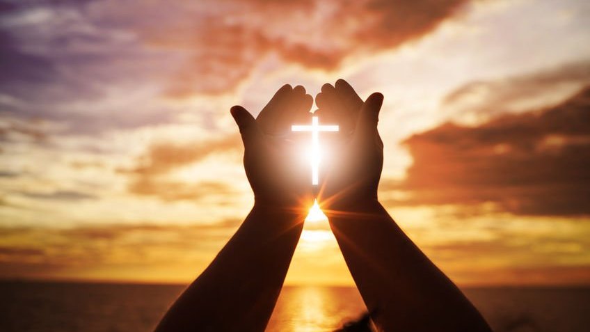 Mãos abertas para adoração com o símbolo da cruz no meio, e no fundo um pôr do sol
