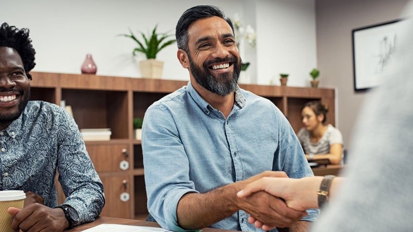 Homem de camisa azul sorrindo dando um aperto de mãos com outra pessoa.