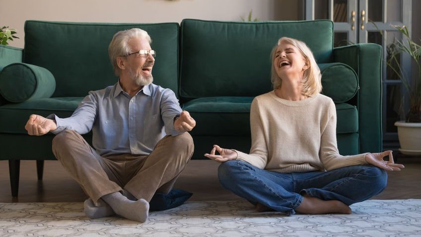 Casal de idosos sorrindo enquanto meditam na sala de estar.