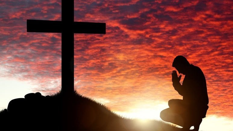 Homem ajoelhado rezando em frente de uma cruz com o pôr do sol de paisagem