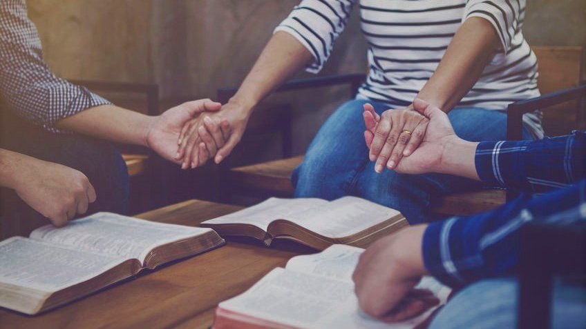 Grupo de três pessoas dando as mãos em uma roda, em volta de uma mesa com três bíblias abertas