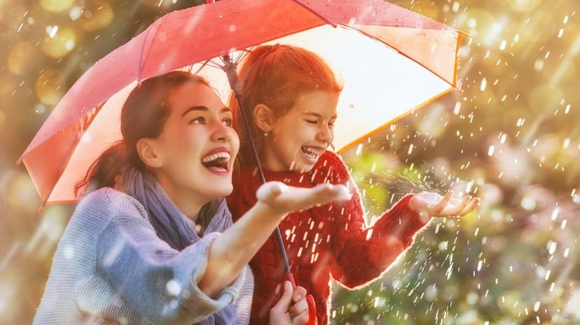 Menina de blusa vermelha e sua mãe felizes embaixo de um guarda-chuva enquanto chove