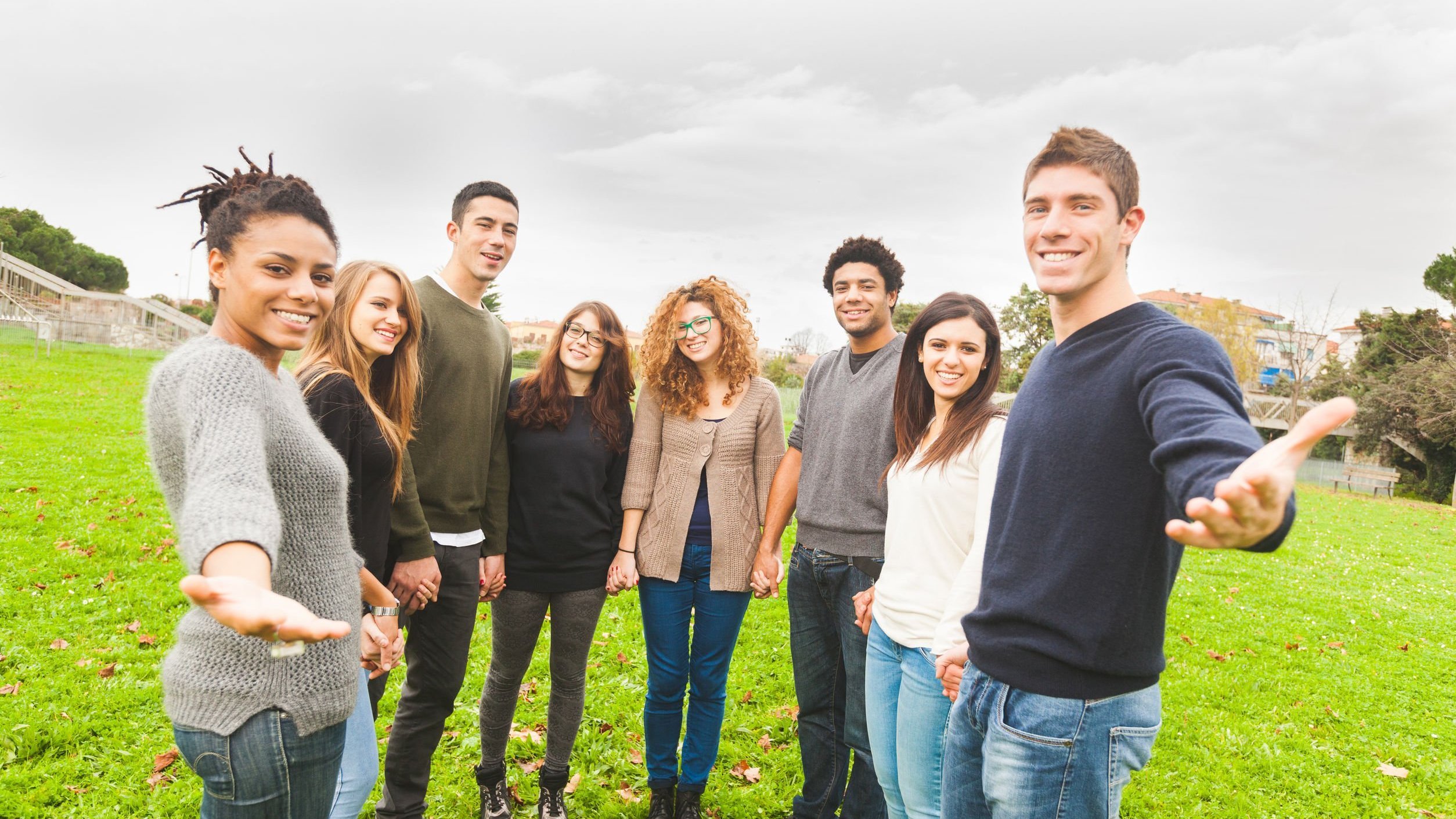 Grupo de amigos sorridentes. Mulher da extremidade esquerda e homem da extremidade direita estendem os braços.