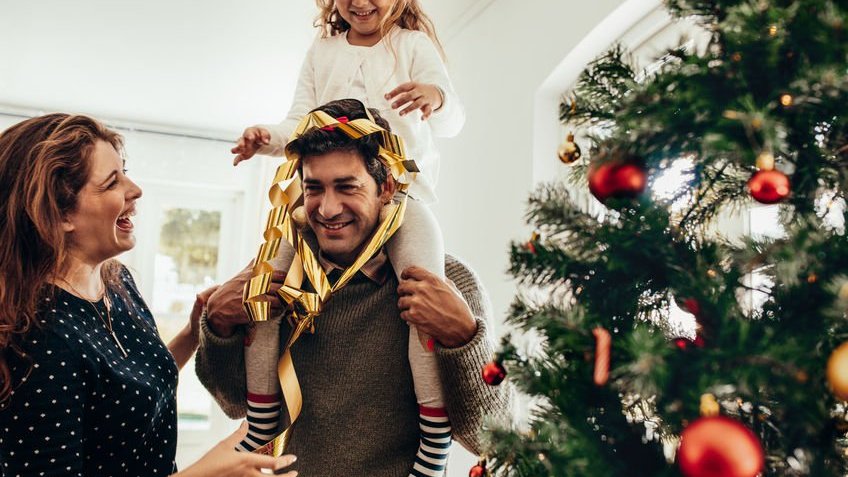 ammentorp / 123RF Uma menina sentada nos ombros de seu pai com uma árvore de natal ao lado.