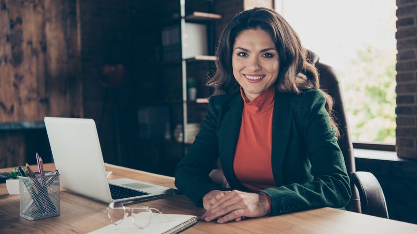 Mulher morena sorrindo sentada em frente uma mesa com computador aberto em um escritório