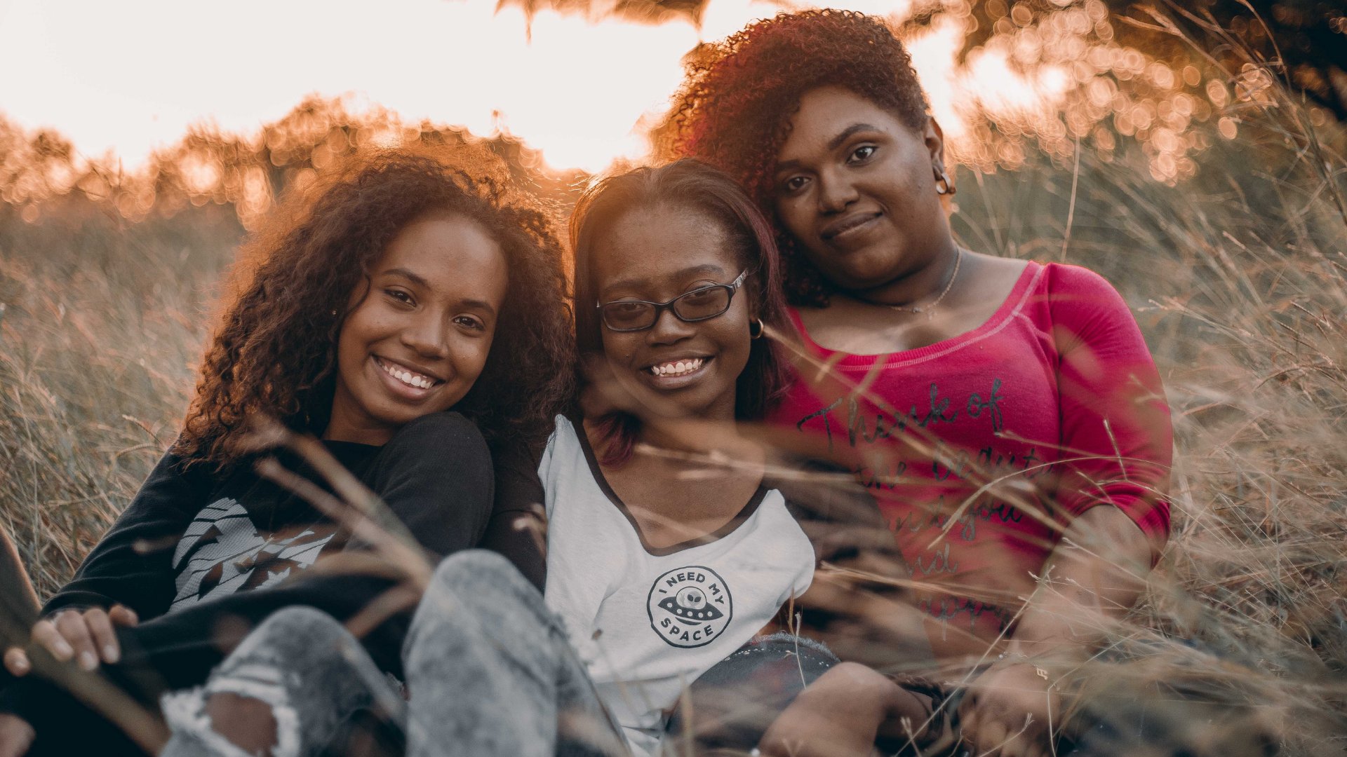 Três mulheres negras sorrindo, enquanto se abraçam, sentadas na grama de folhagem alta e com um pôr do sol ao fundo