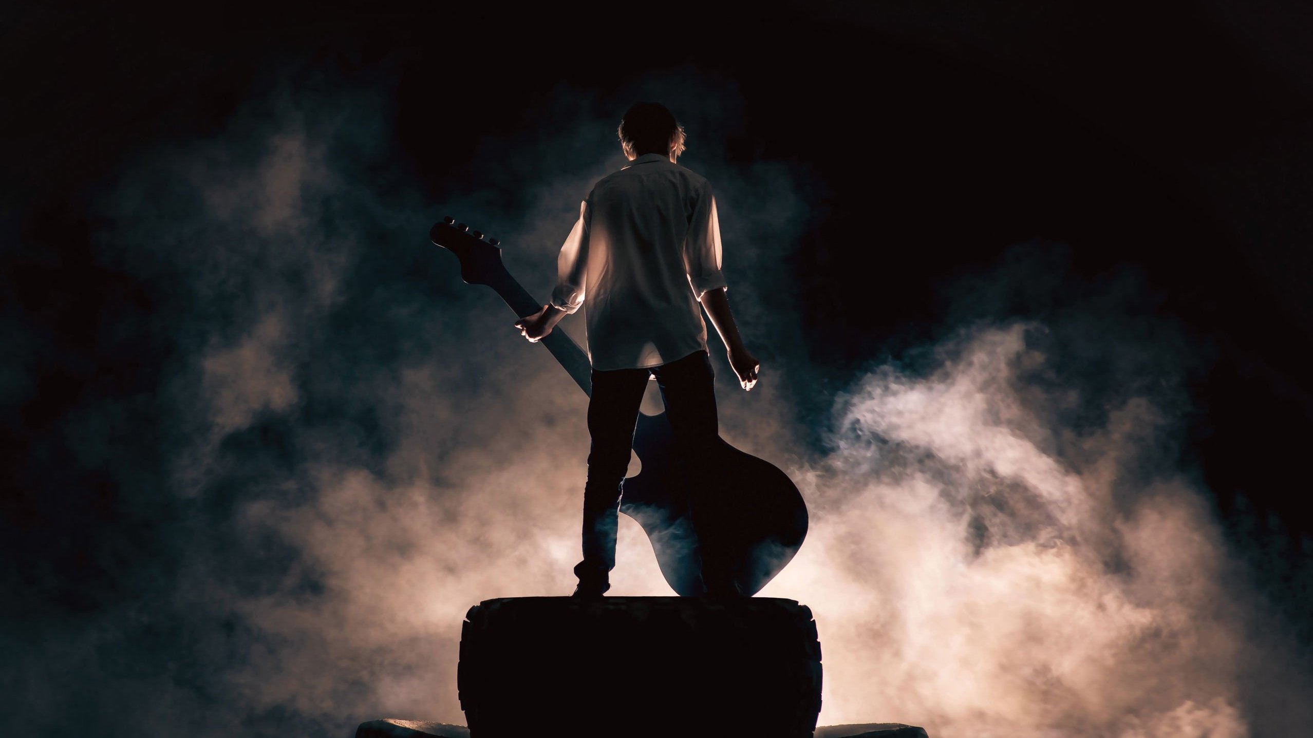 Homem segurando guitarra, rodeado de fumaça, em cima de palco de show